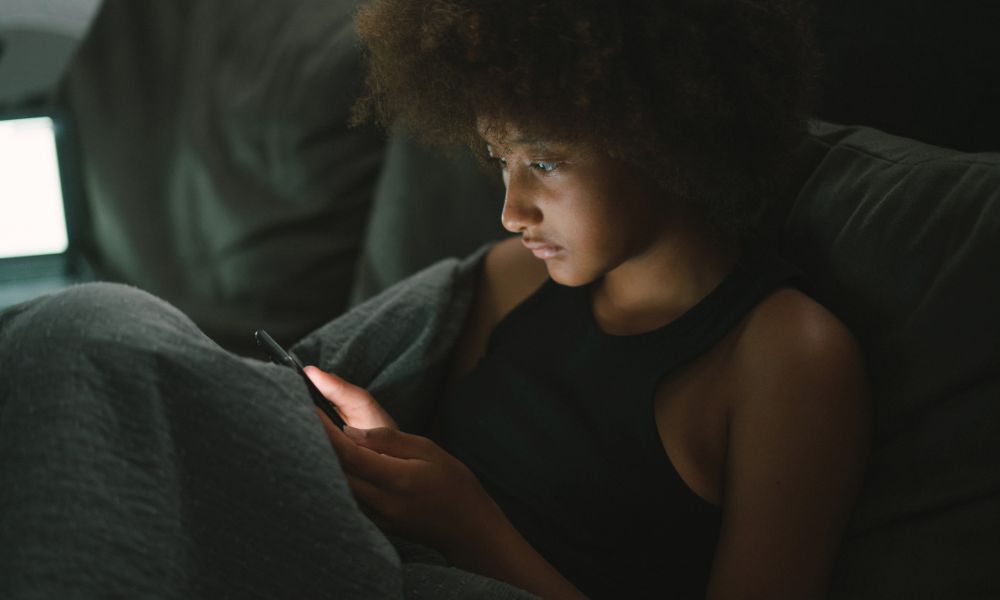 Teen sitting in a dark room, wrapped in a blanket and looking intently at a smartphone, reflecting stress, isolation, or emotional processing—illustrating the mental challenges that can shape resilience during adolescence.