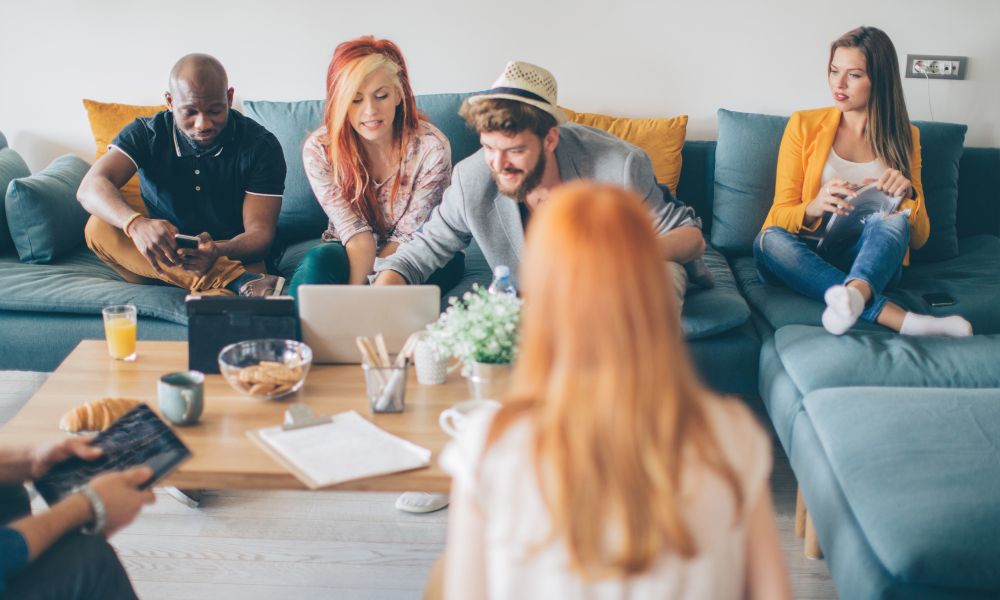 Group of young adults gathered in a casual living room setting, talking, using laptops and tablets, and engaging in conversation—representing the power of social connection and peer support in building resilience.