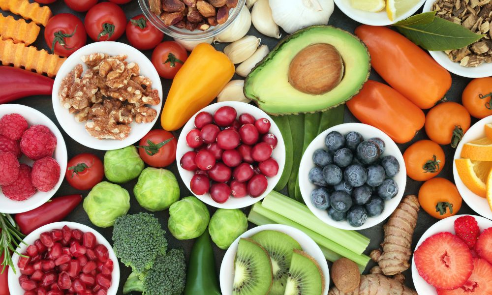 Colorful assortment of fresh fruits, vegetables, nuts, and herbs arranged on a table, including blueberries, raspberries, avocado, oranges, kiwi, tomatoes, peppers, broccoli, walnuts, and garlic, representing healthy foods that support gut and brain health.