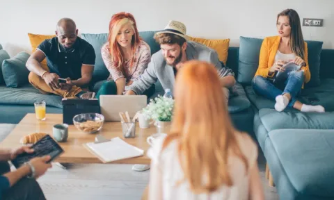 Group of young adults gathered in a casual living room setting, talking, using laptops and tablets, and engaging in conversation—representing the power of social connection and peer support in building resilience.