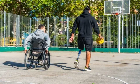 Two men enjoy time together on an outdoor basketball court. One man uses a wheelchair and the other has a prosthetic leg and is holding a basketball. Trees and a basketball hoop are visible in the background.