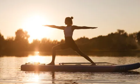 Woman practicing yoga on a paddleboard at sunrise, standing in warrior pose on calm water with trees in the background, symbolizing balance, resilience, and mental strength.