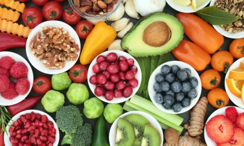 Colorful assortment of fresh fruits, vegetables, nuts, and herbs arranged on a table, including blueberries, raspberries, avocado, oranges, kiwi, tomatoes, peppers, broccoli, walnuts, and garlic, representing healthy foods that support gut and brain health.